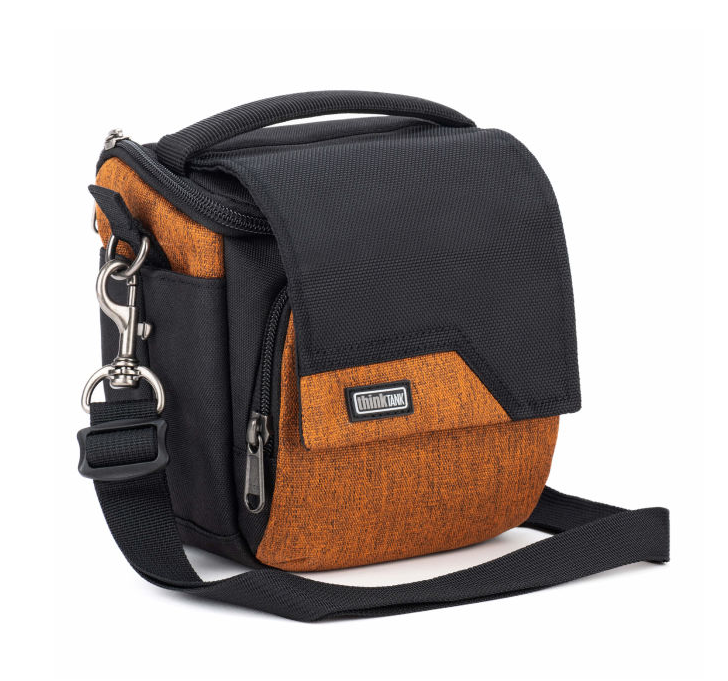 A small camera bag with a black nylon body and a textured orange-brown fabric front pocket, featuring a top handle, front zipper, side pocket, and adjustable shoulder strap on a white background.